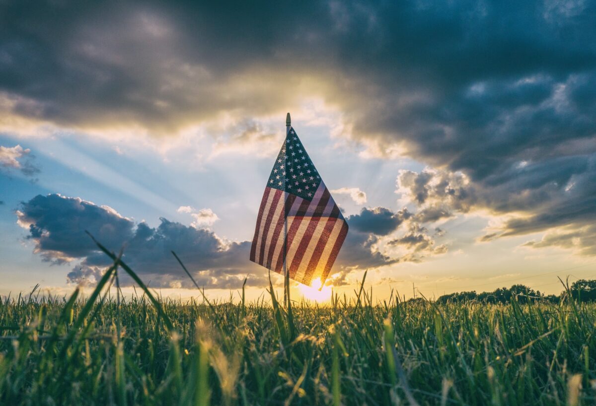 US flag in a green field with clouds & sunlight in the sky behind the flag