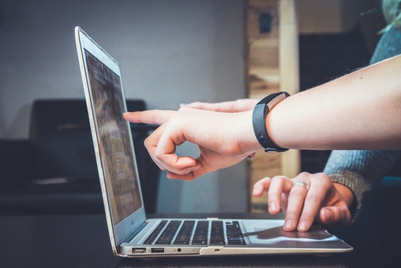 person's hand pointing at a laptop screen with another hand touching the keyboard